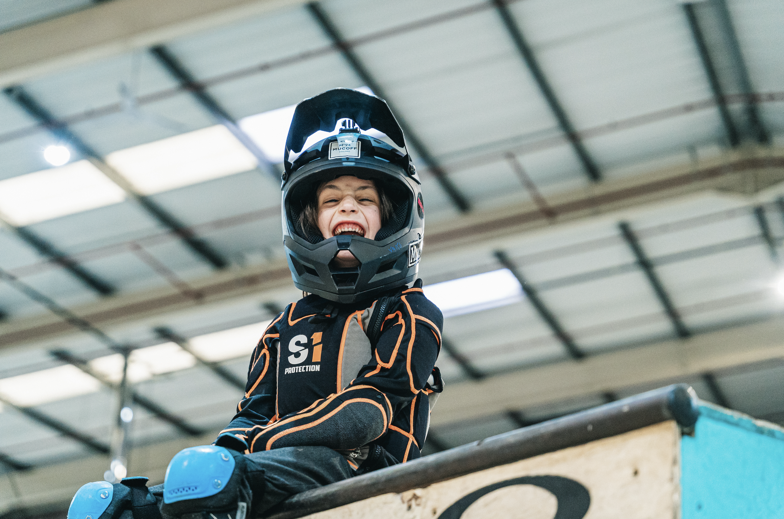 A young athlete smiling with his mouth open, wearing a full-face helmet and protective gear, sitting on a ramp in an indoor skate park for a shoot for British Cycling and Paris Olympics