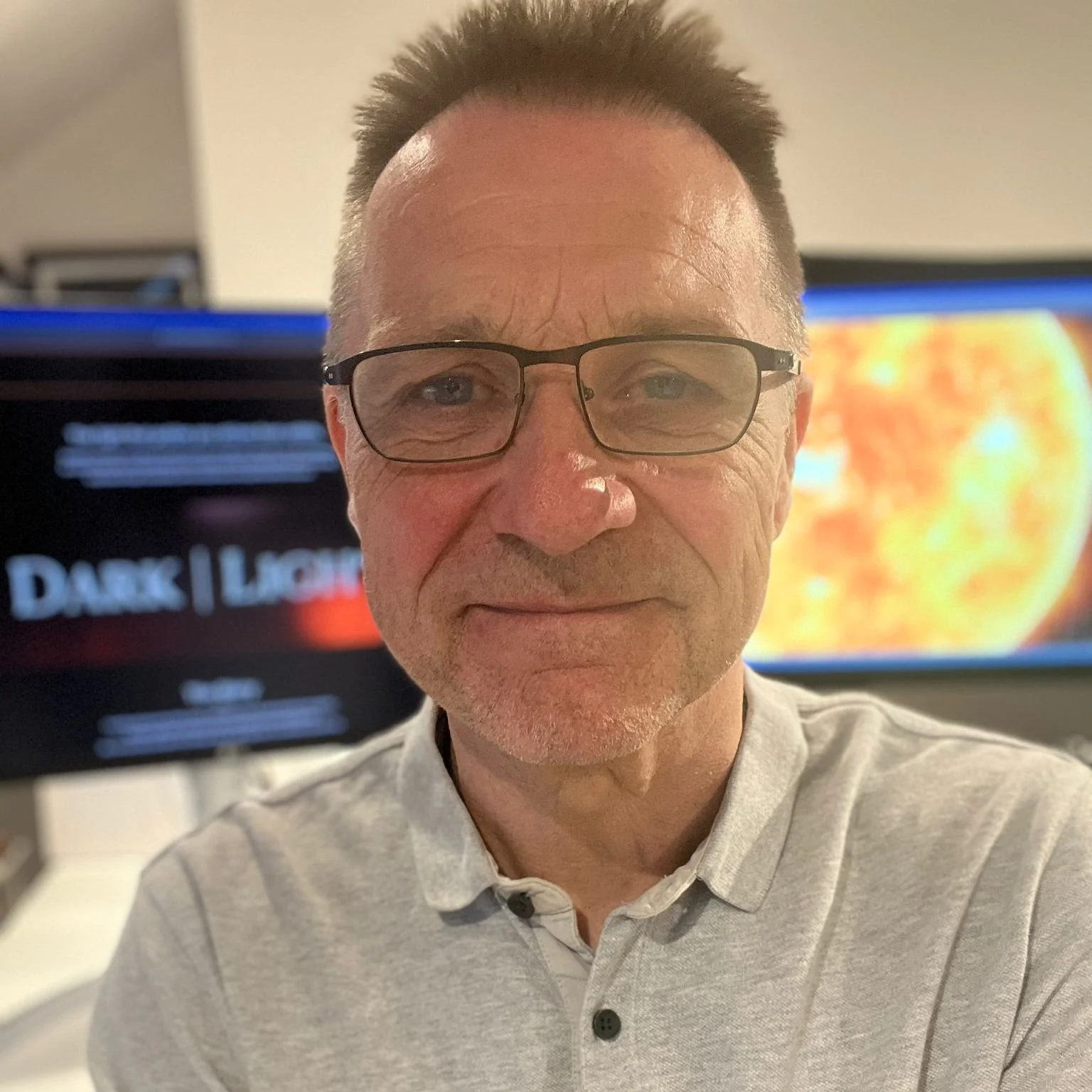 Head of Production man with glasses, smiling, wearing a light gray polo shirt, with post production screens in the background.