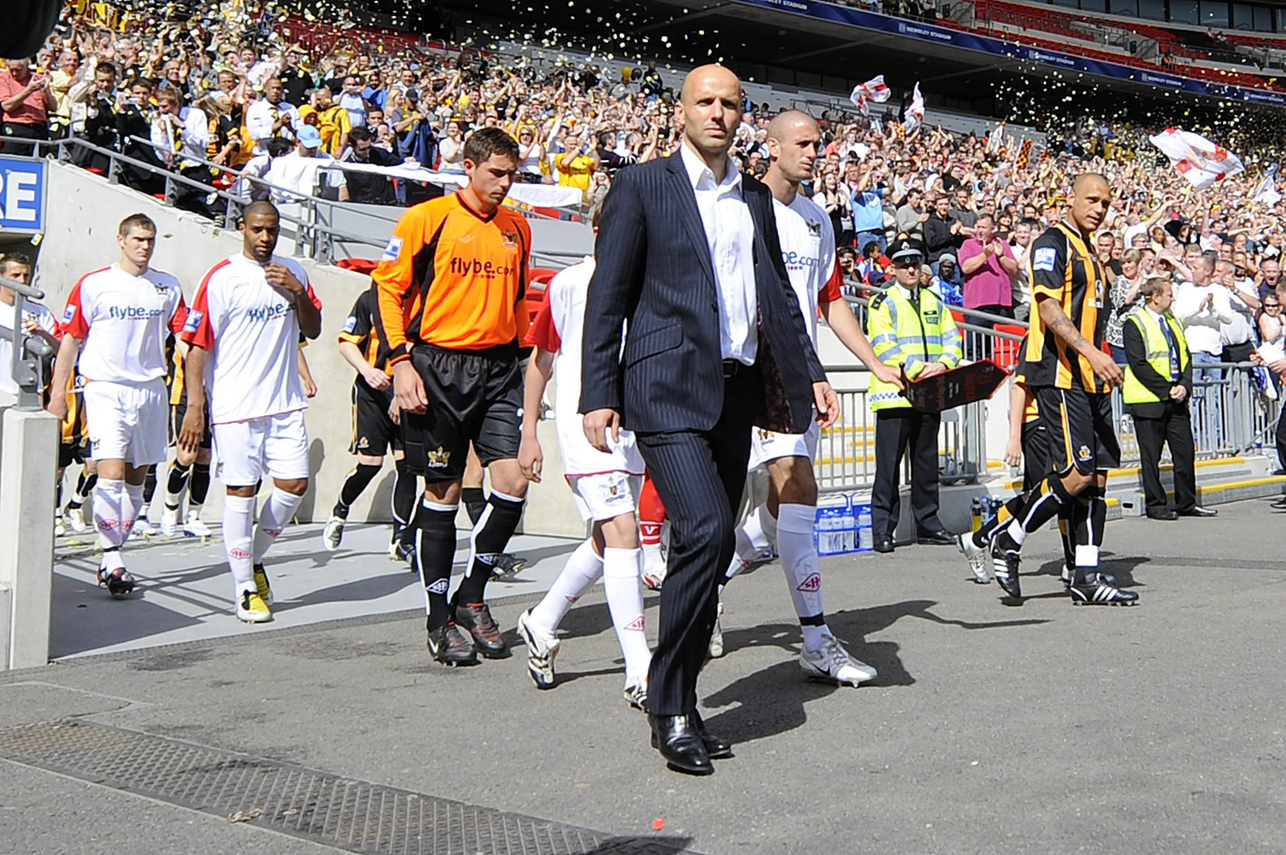 Soccer players and team officials walking onto the field with a large crowd in the stadium stands cheering in the background.