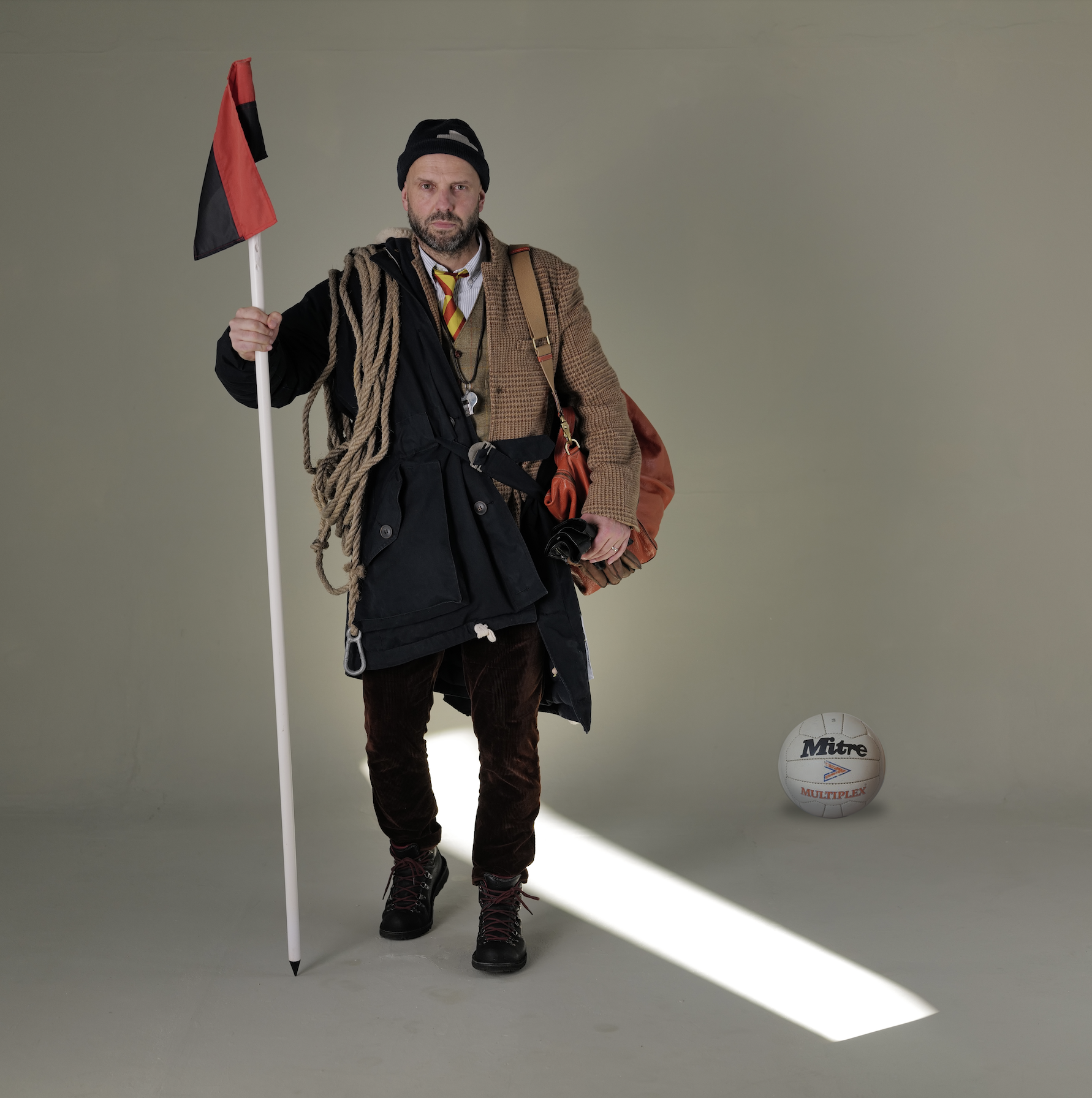 A man dressed in outdoor hiking gear holding a flag stands in a studio with a volleyball on the floor to his right. The background is plain and neutral.