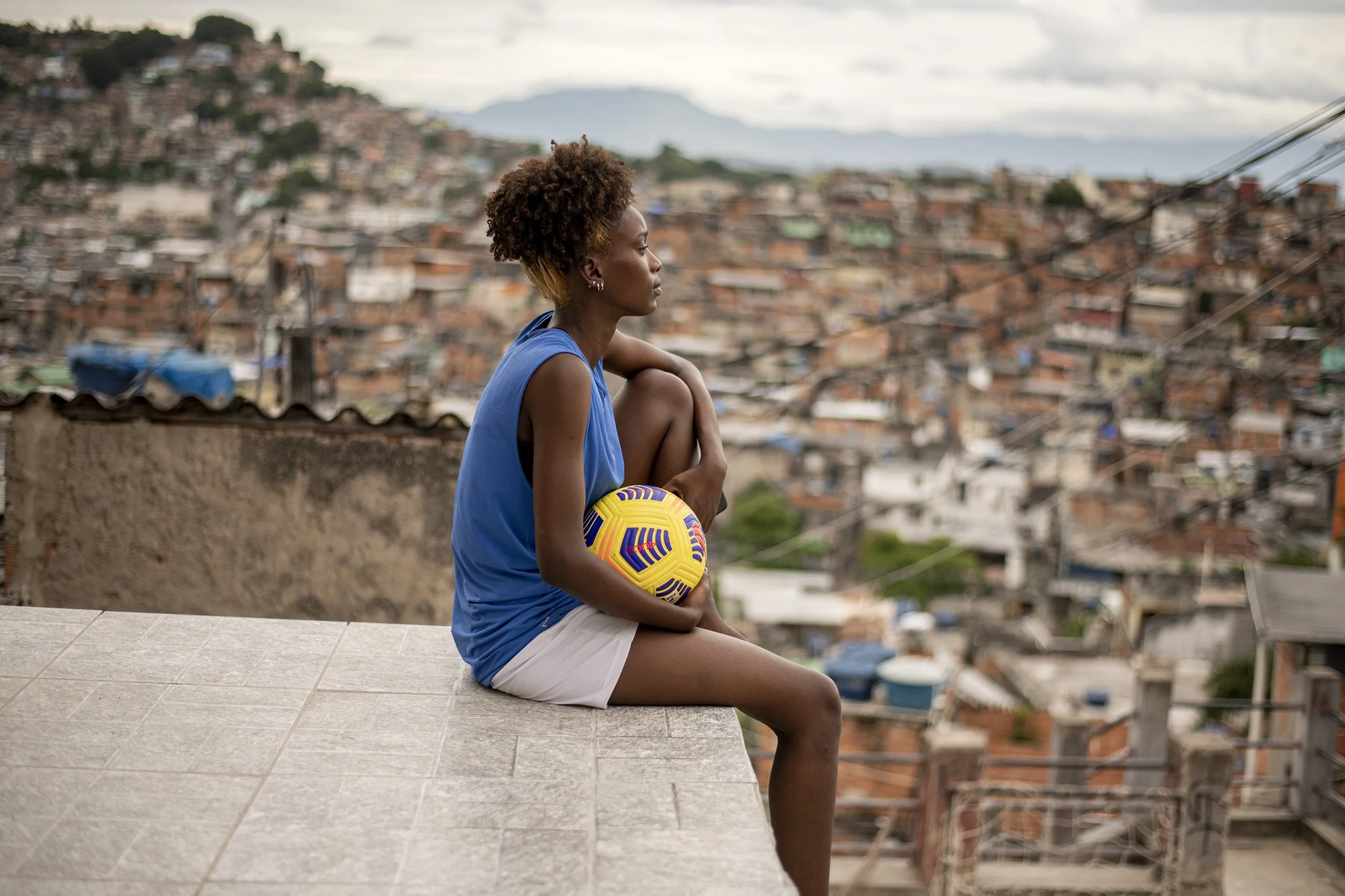 A woman in a blue sleeveless shirt and white shorts sitting on a concrete ledge, holding a colorful soccer ball, with a cityscape of densely packed houses on a hillside in the background.