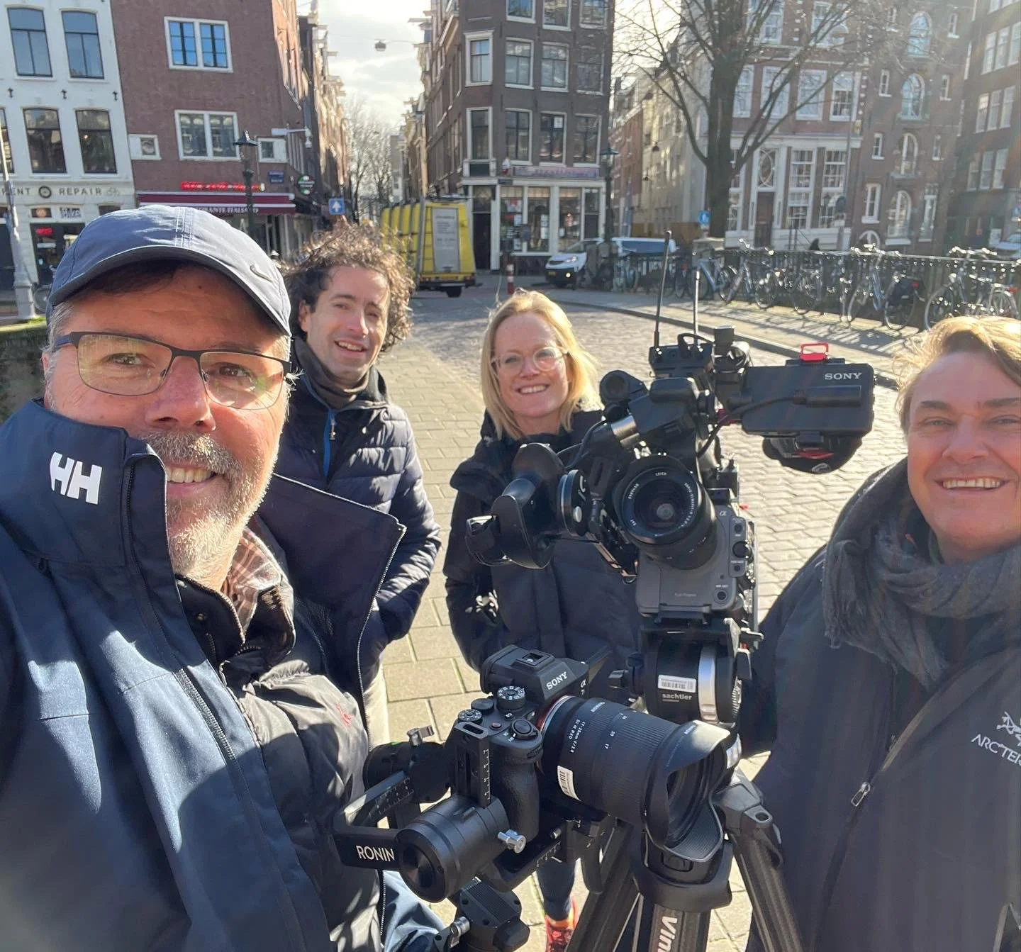Four smiling people, two men and two women, taking a selfie outdoors on a sunny day. They are standing behind professional video camera equipment on a city street with brick buildings, parked bicycles, and trees in the background.