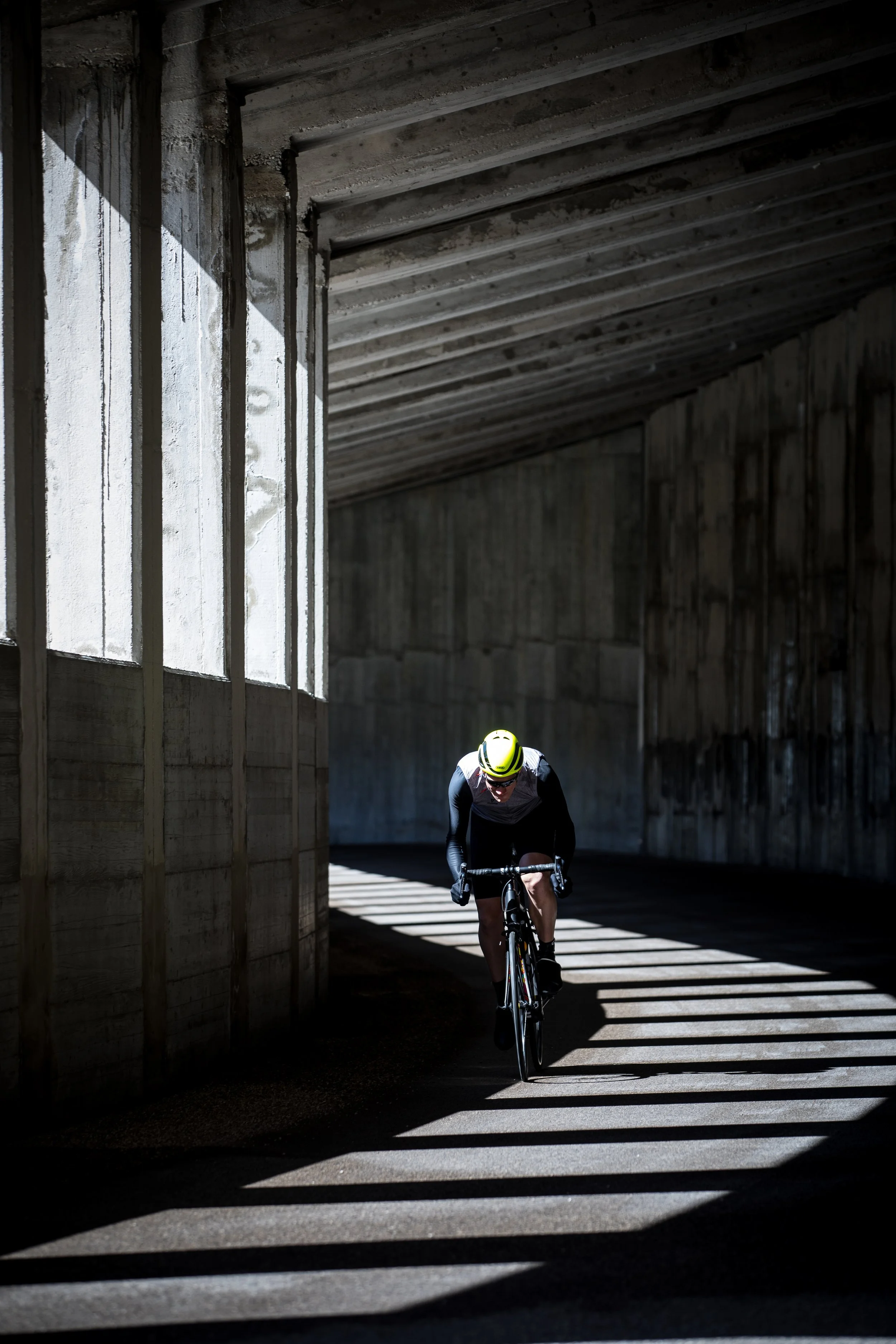 Male cyclist riding under a concrete bridge with sunlight creating striped shadows on the ground.