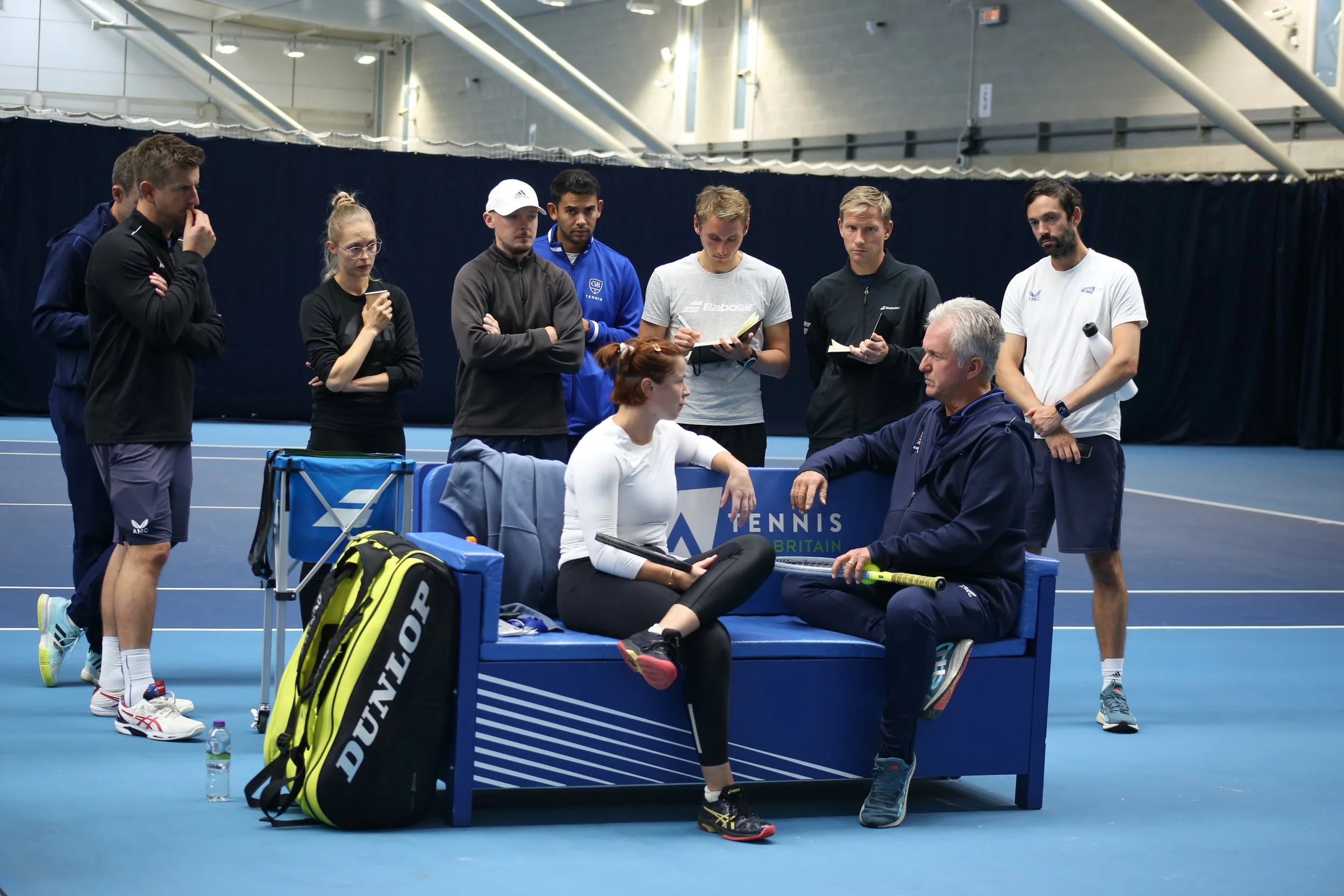 A female tennis player sitting on a blue bench in an indoor tennis court, talking to her coach who is sitting beside her. Several people, including coaches and staff, are standing behind them, taking notes or observing, with one person holding a tennis racket. Tennis equipment is visible, including a tennis bag and water bottles.