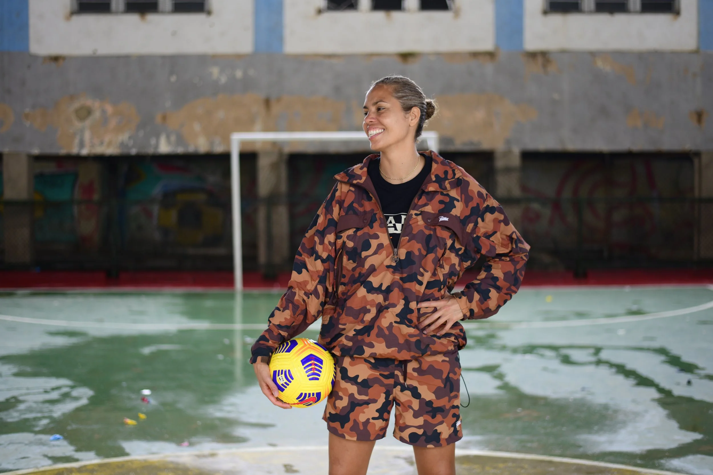 Woman in camouflage sportswear holding a yellow soccer ball stands on an outdoor sports court.