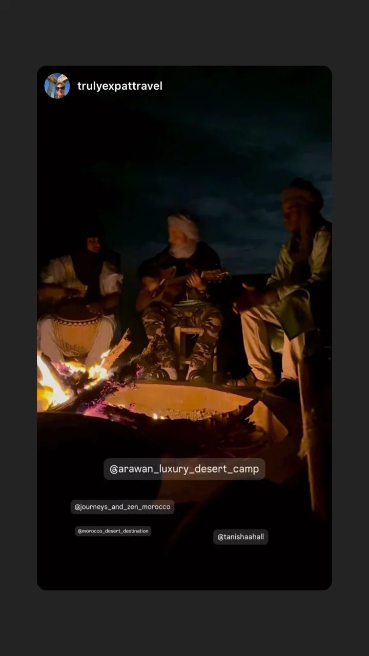 Group of people sitting around a campfire at night, some playing guitars and singing, in a desert setting in Morocco.