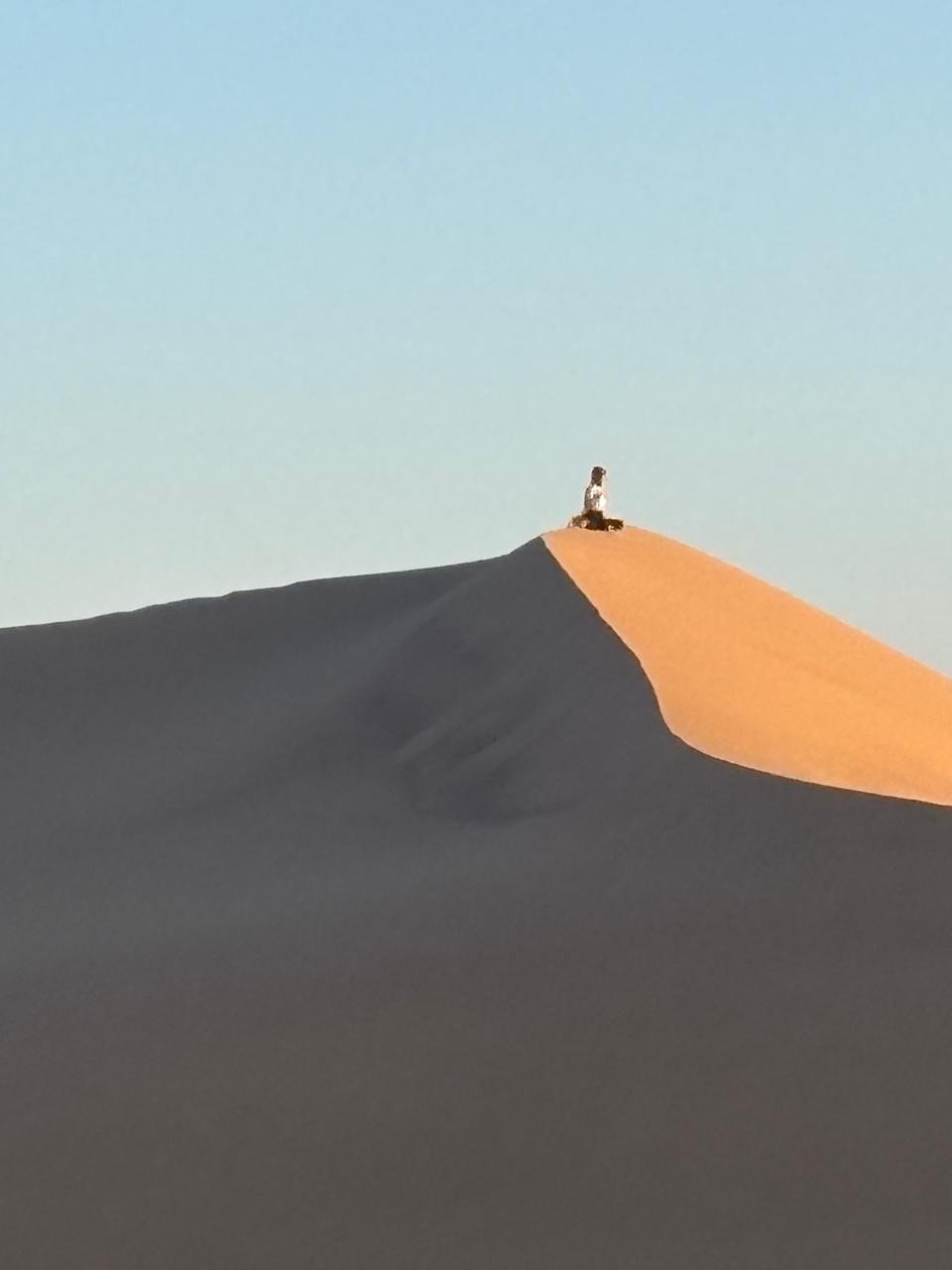 A person sitting at the top of a sand dune in a desert, with sunlight illuminating the crest, creating contrasting shadowed and lit areas on the dunes.
