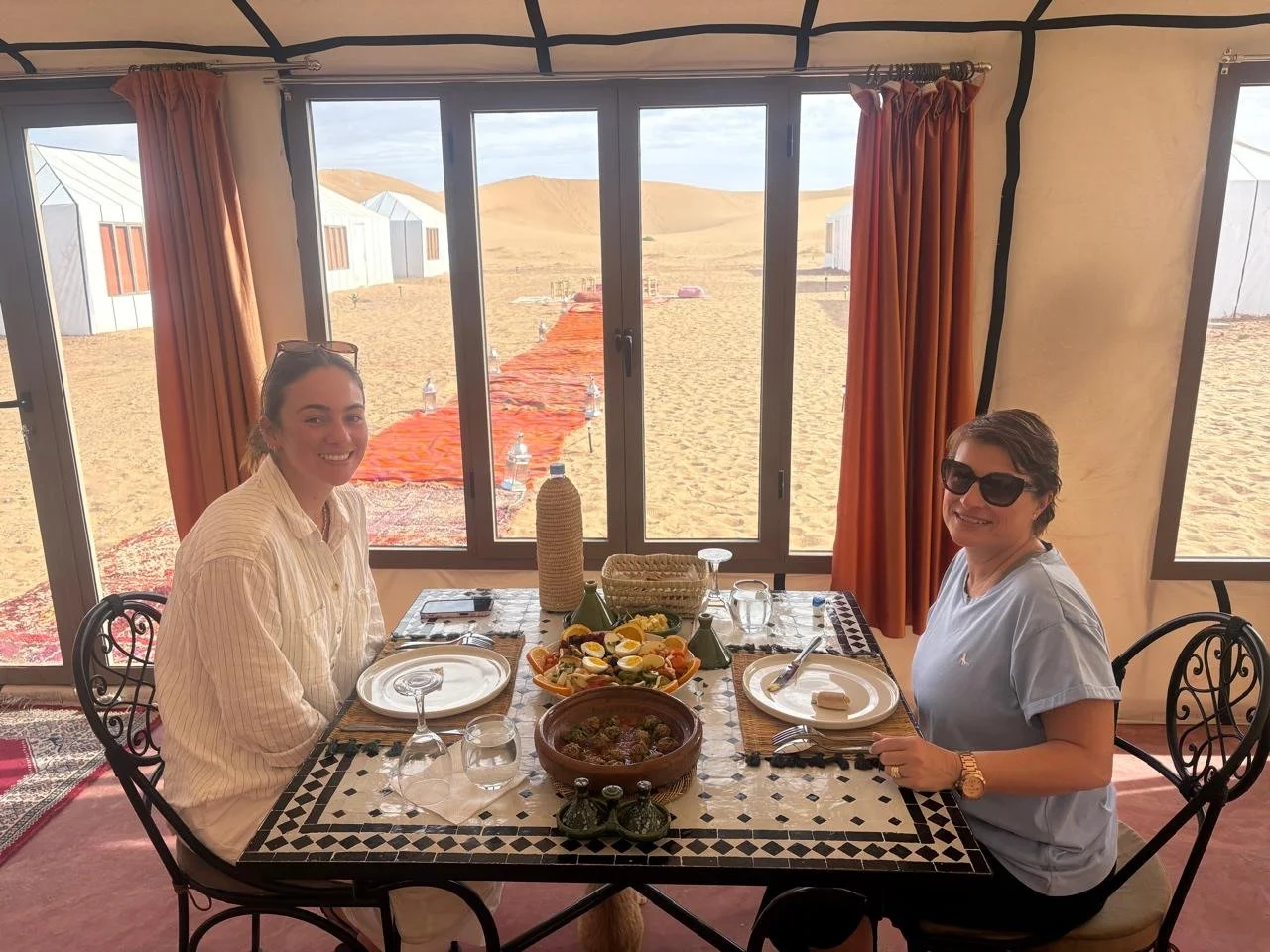 Two women sitting at a dining table inside a desert tent with a view of sand dunes through large windows. The table has plates, glasses, and a variety of food, including a salad, a dish in a clay pot, and a bottle.
