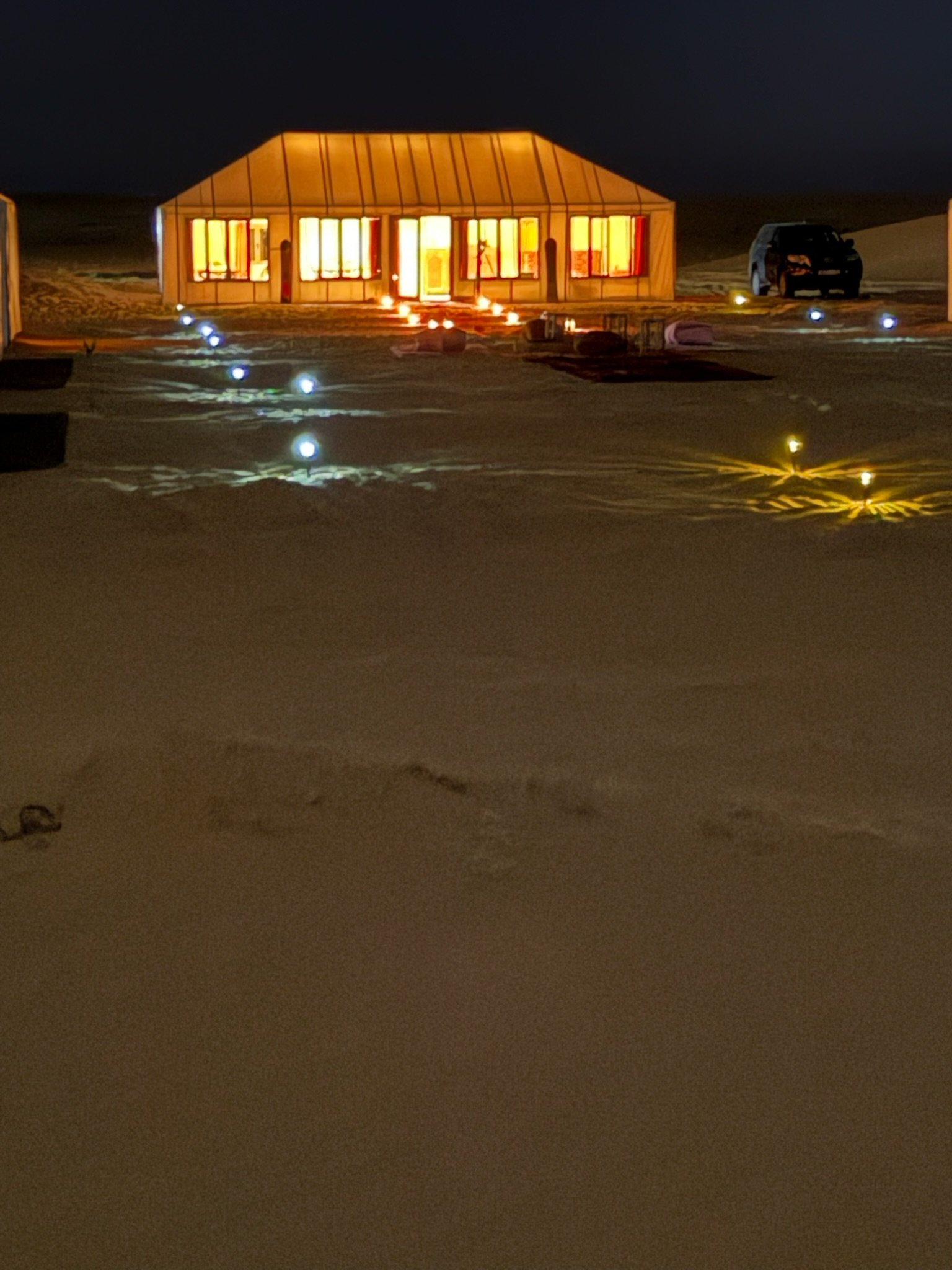 Nighttime view of a lit-up tent or house with illuminated pathways and a vehicle in the background.