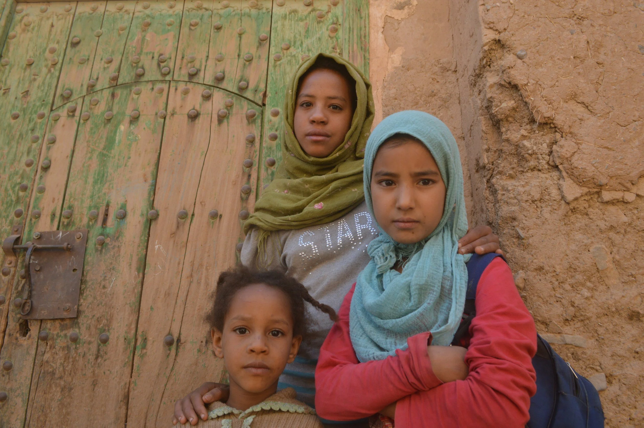Three children standing in front of a rustic wooden door with green paint and metal embellishments, and a rough brick wall. They are wearing scarves and casual clothing.