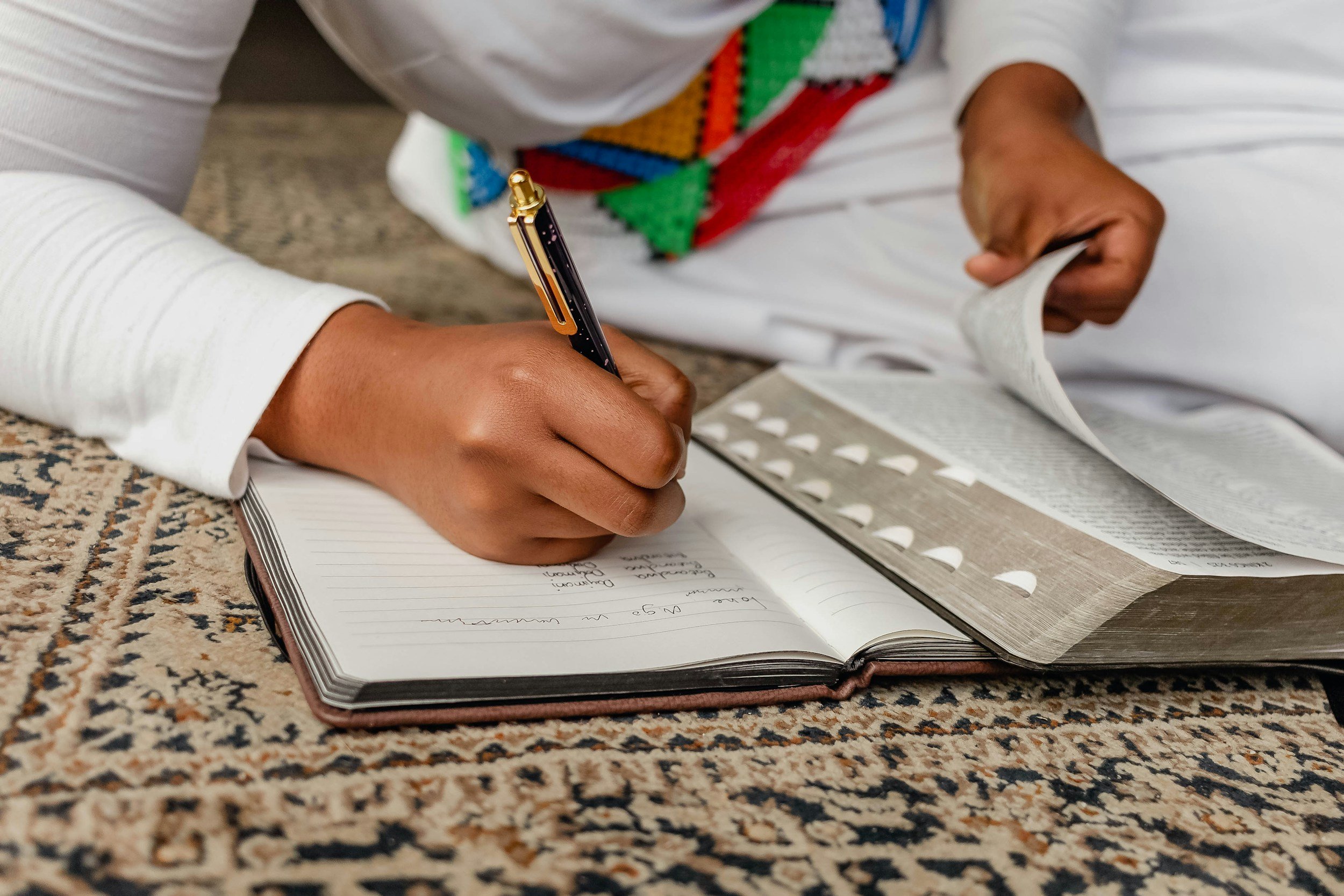 A person wearing a white shirt is writing in a notebook with a pen, while holding a newspaper on a textured surface.