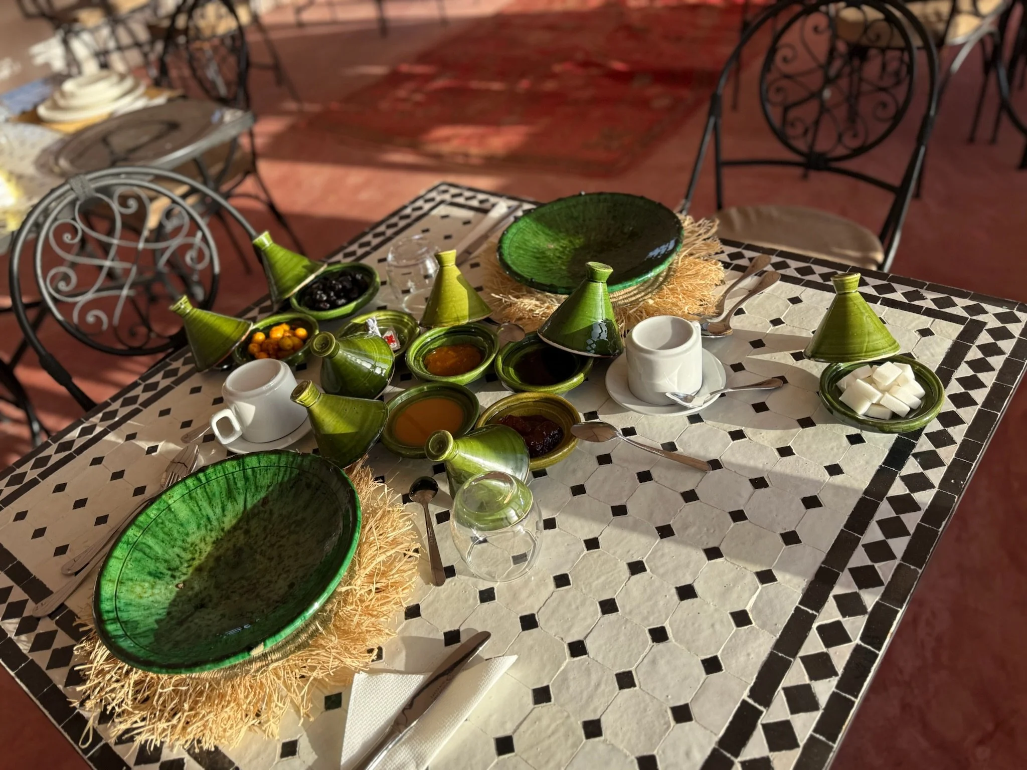 A table set for a meal with green ceramic dishes, small bowls of condiments, cups, glasses, and cutlery on a white and black patterned tablecloth.