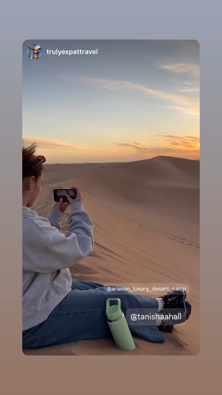 Person sitting on desert sand dunes during sunset, taking a picture with a smartphone, with a green water bottle beside them.