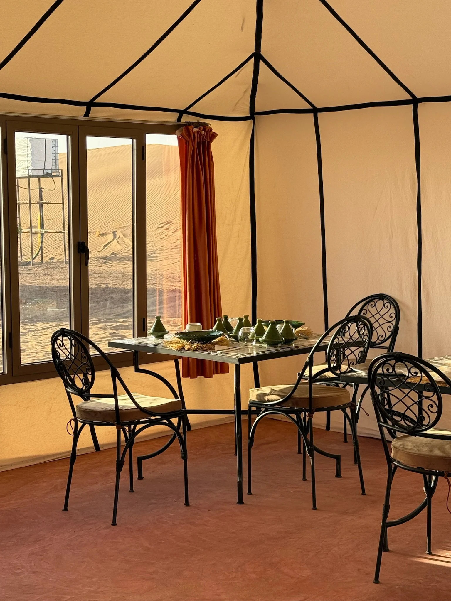 Interior of a tent with a dining table set with green dinnerware and four black wrought iron chairs, with a view of sand dunes outside through sliding glass doors.