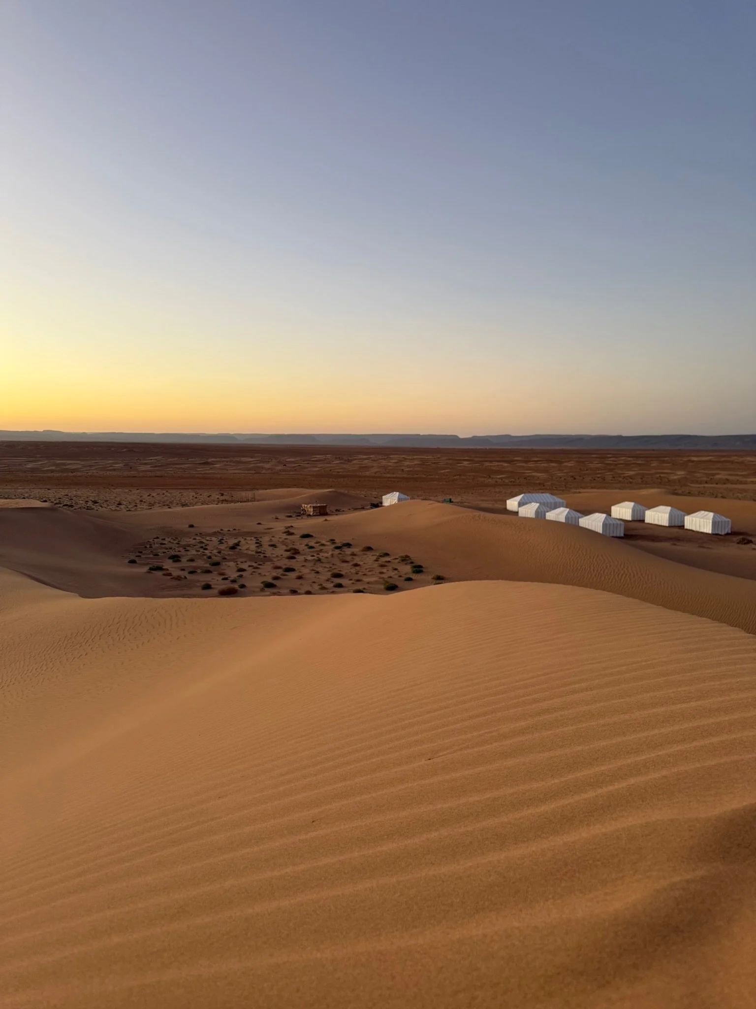 Desert landscape at sunset with sand dunes and white tents in the distance.