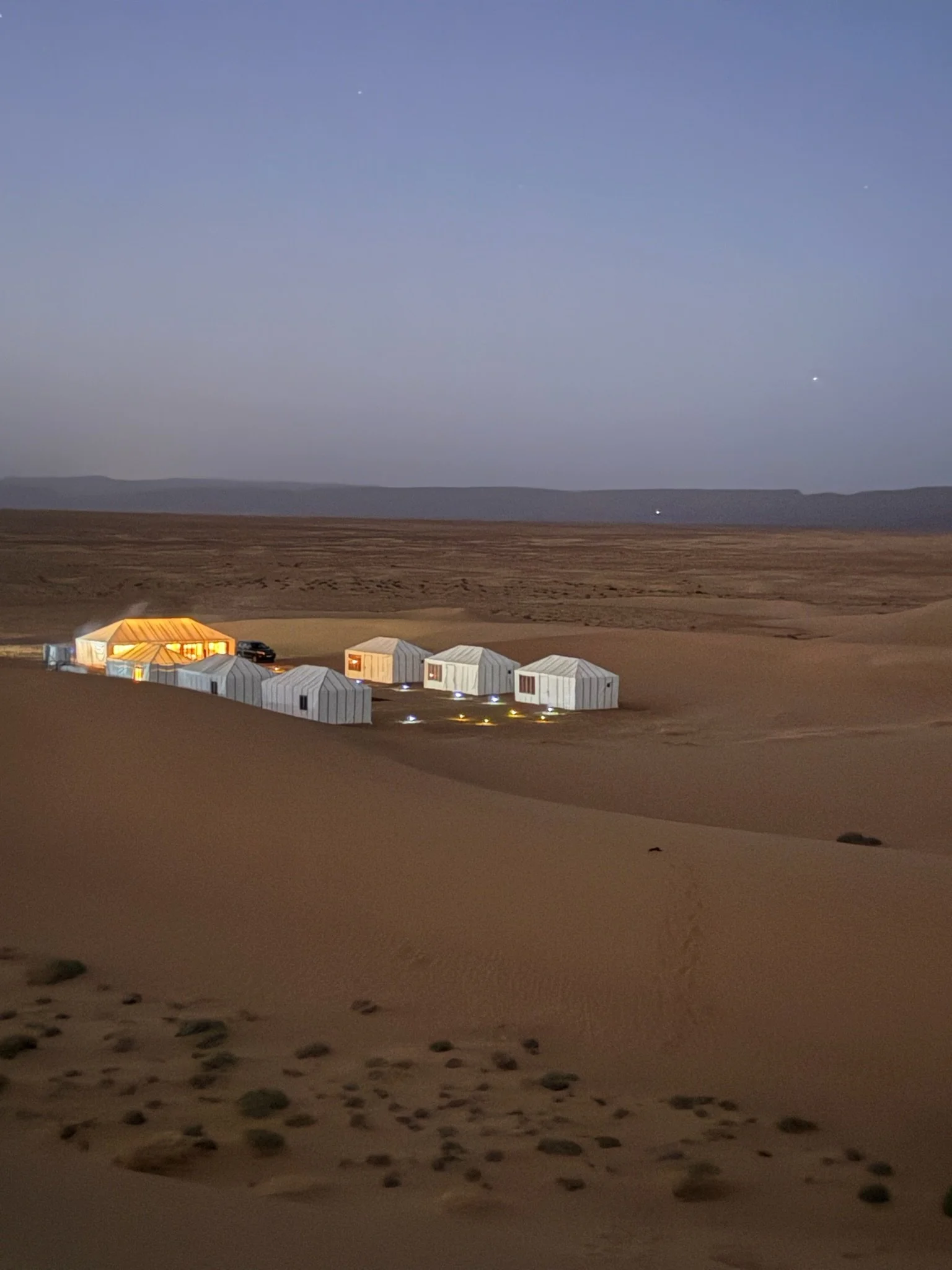 Several white tents and a large tent illuminated with warm light in a desert at dusk, with a few vehicles parked nearby and a flat, sandy landscape extending to distant mountains under a twilight sky.