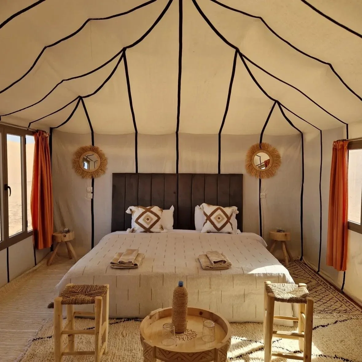 Bedroom inside a tent with a bed, pillows, bedside tables, mirrors, and curtains, with black lines outlining the tent's interior.