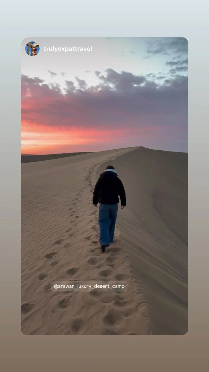 Person walking on sand dunes during sunset or sunrise with footprints behind, in a desert setting, tagged with @arawan_luxury_desert_camp.