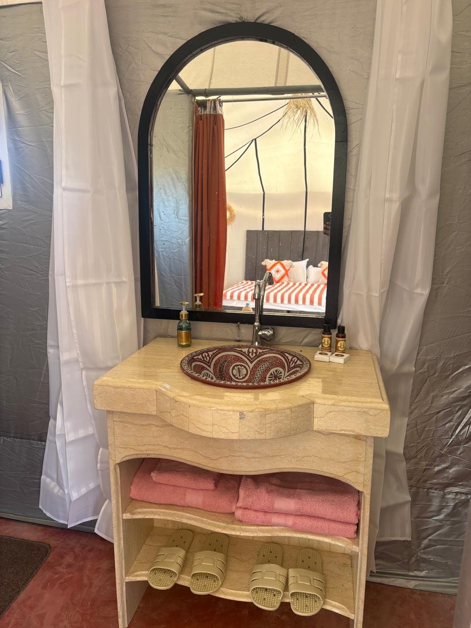 A bathroom vanity with a decorative bowl sink, two pink towels, and pairs of slippers underneath. A large mirror is mounted above the sink, reflecting a bedroom with a bed that has a striped bedspread and orange pillows. The vanity also has soap disp