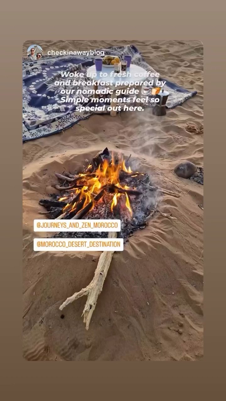 Campfire on sandy ground with a blanket set up nearby, breakfast spread with drinks and food on the blanket, desert landscape in the background.