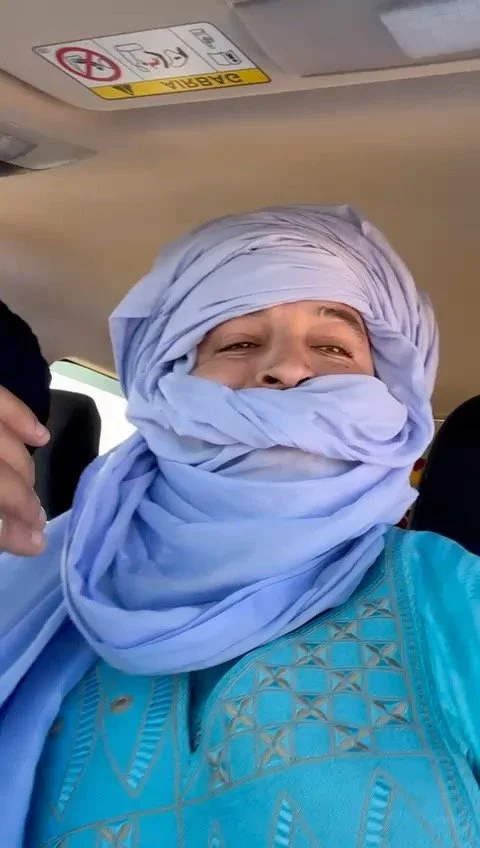 A smiling woman wrapped in a light blue scarf sitting inside a vehicle, with a beige interior and a sign on the ceiling.