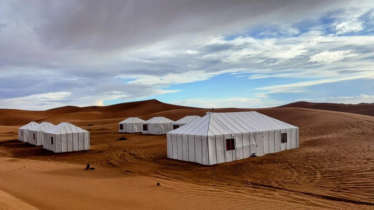Multiple white tents set up in a desert landscape with sand dunes and a cloudy sky above.