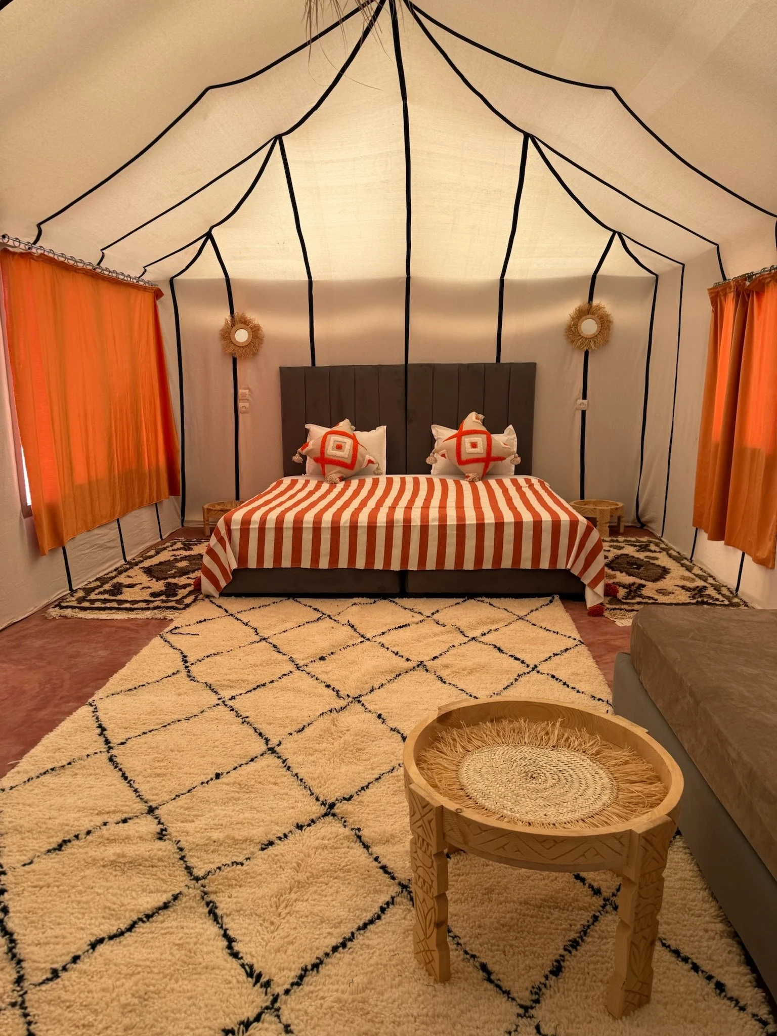 A cozy bedroom inside a tent with orange curtains, a black headboard, and a striped bedspread. The room features two patterned pillows, wall decorations, a large cream rug with black lines, and a small wooden table.