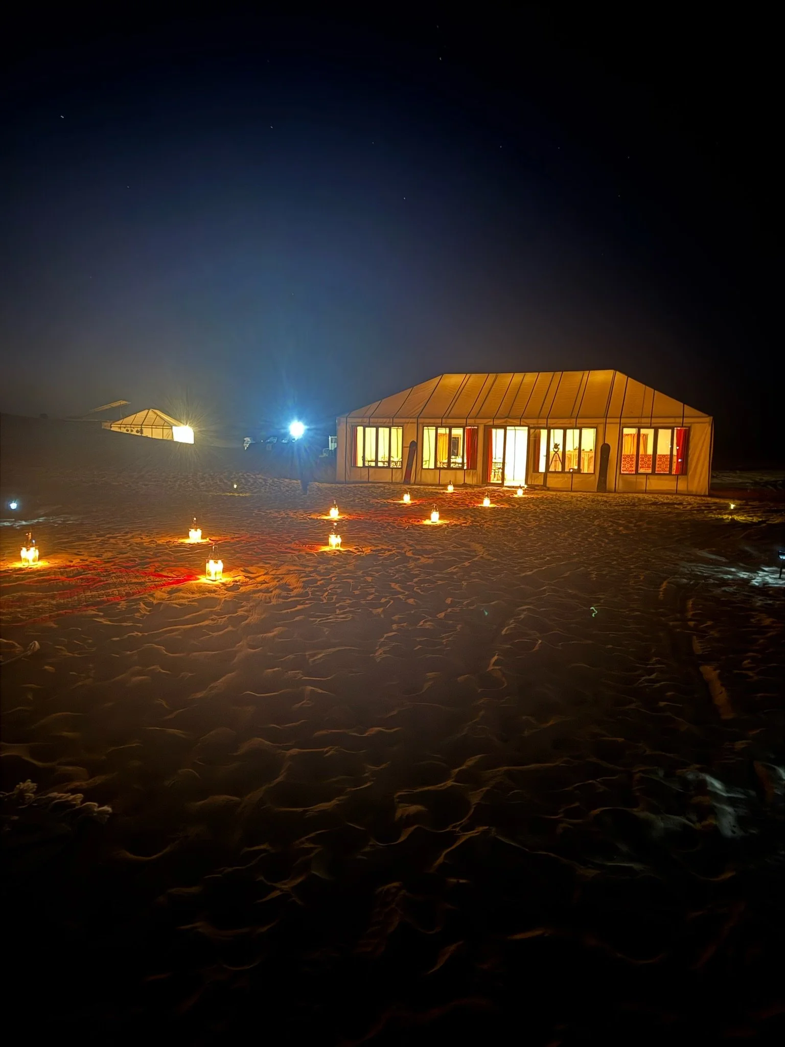 A lit tent on sand at night with candles arranged in a line leading to it, under a starry sky.