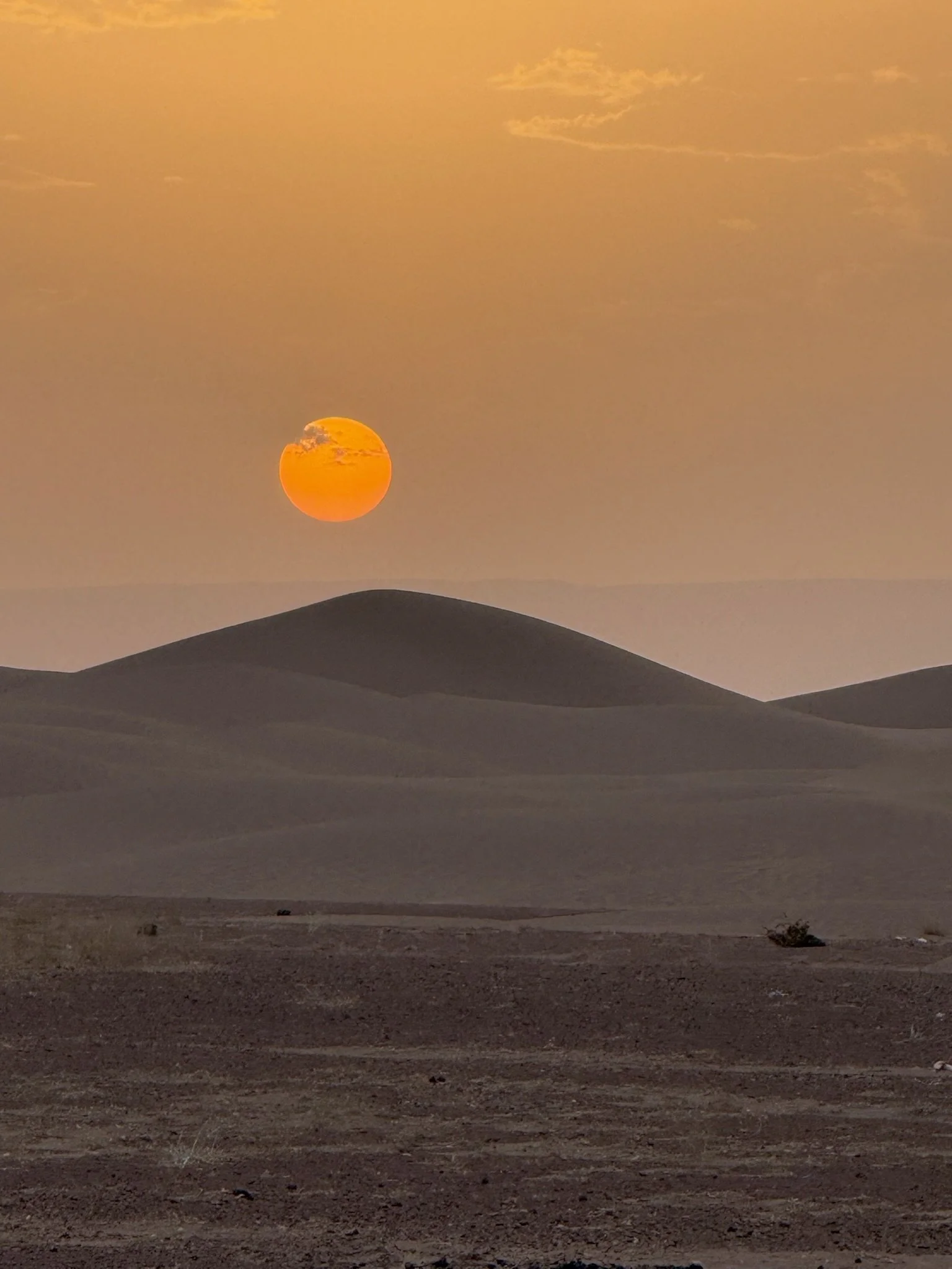 Sunset in a desert with sand dunes and a low sun in the sky, partly covered by clouds.