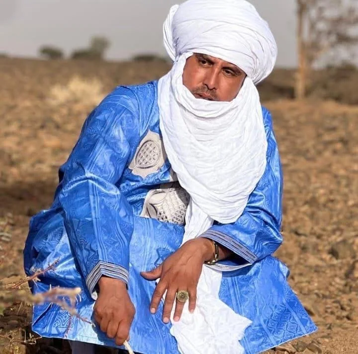 A man in traditional attire with a white turban and blue robe, sitting outdoors on a dirt ground with a neutral expression.