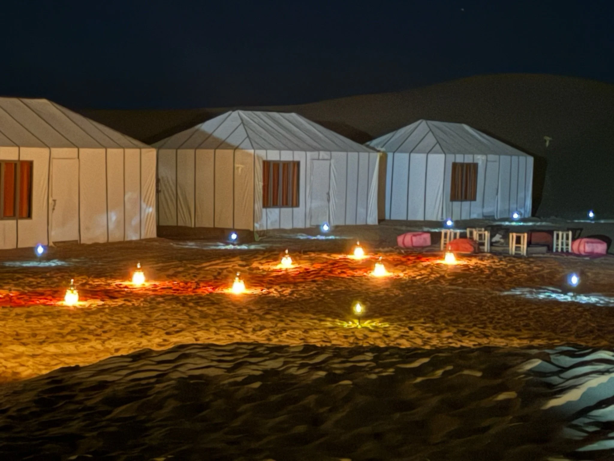 A nighttime scene of a desert with three white tents, decorated with small lights, and pink lounge chairs and tables outside.