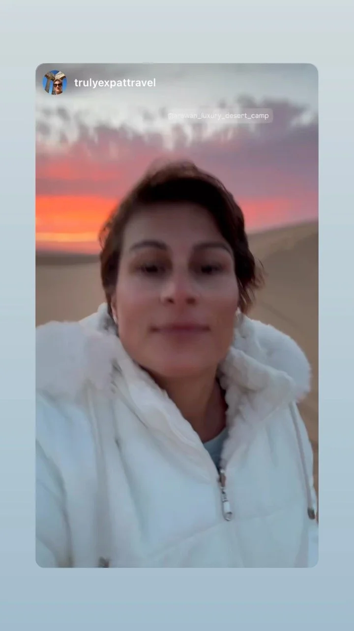 Woman taking a selfie outdoors at sunset, wearing a white jacket in a desert landscape.