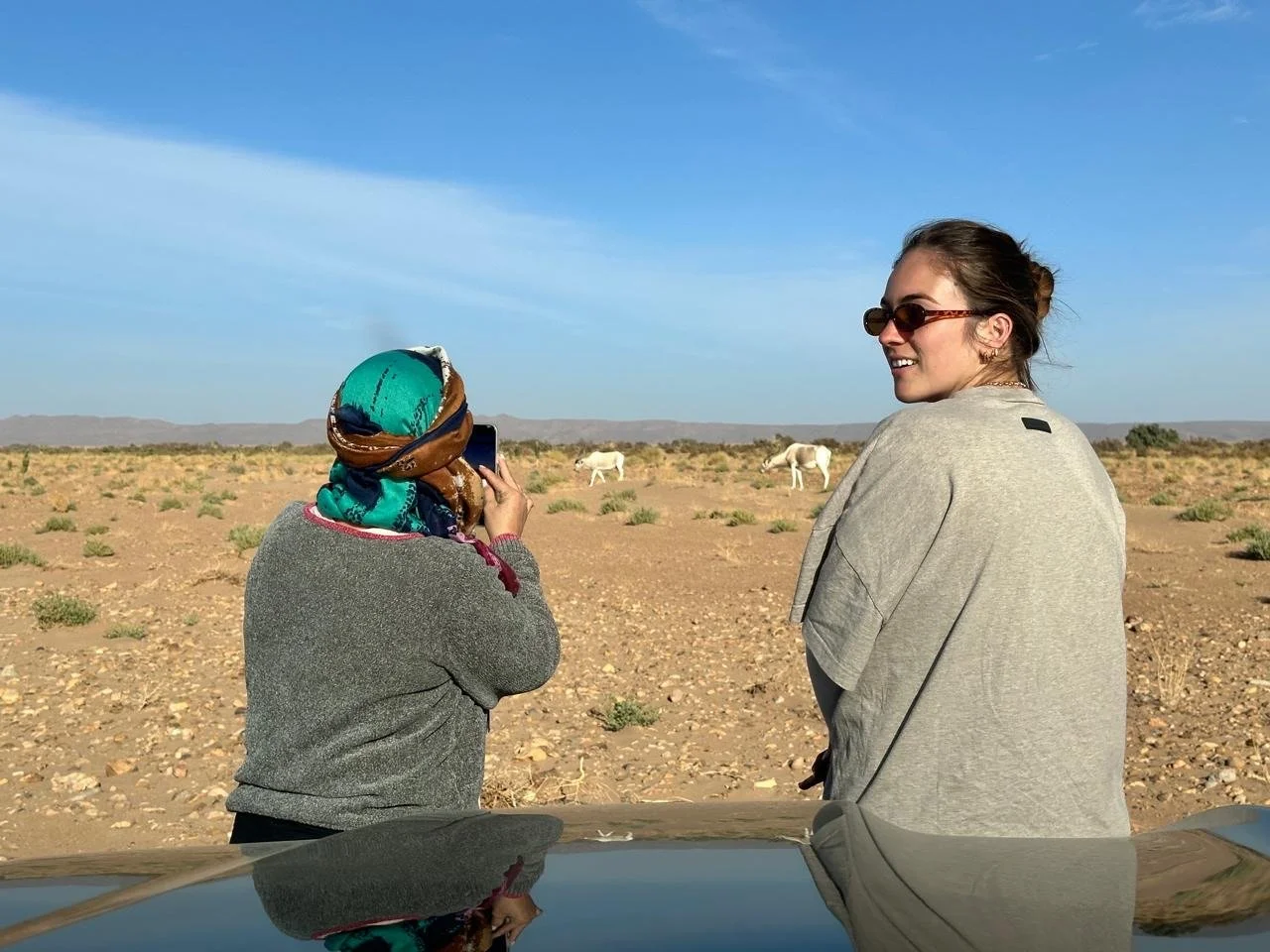 Two women standing outdoors in a desert landscape; one woman is wearing sunglasses and smiling, the other is wearing a headscarf and taking a photo with a smartphone; horses are visible in the background under a blue sky.