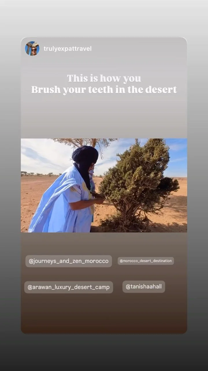 A person brushing their teeth in a desert landscape next to a bush, dressed in traditional white clothing and a headscarf, with blue sky and scattered clouds in the background.
