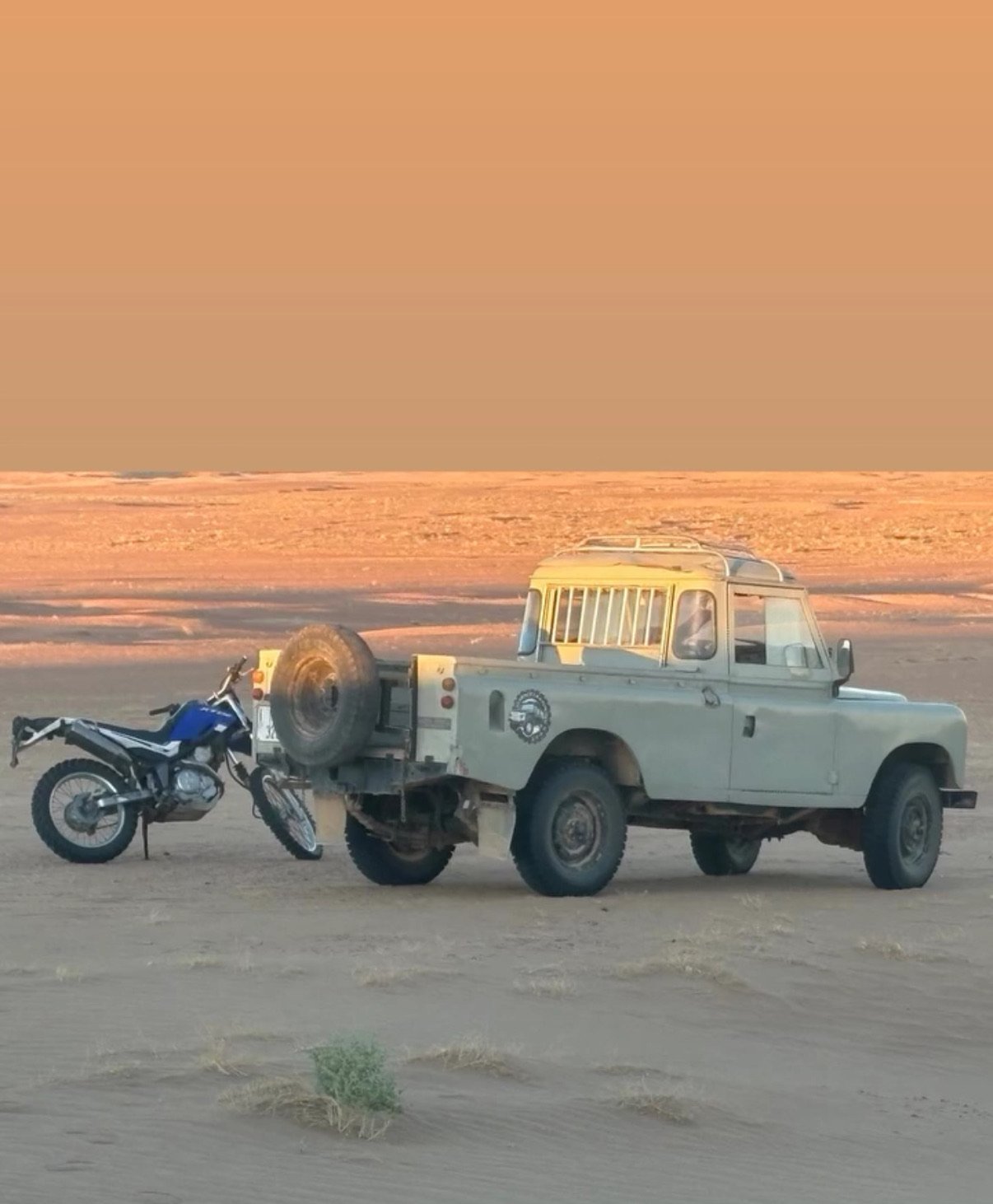 An off-road tan vehicle with a spare tire mounted on the back, parked on the desert, with a blue motorcycle attached to its rear, under a sunset sky.
