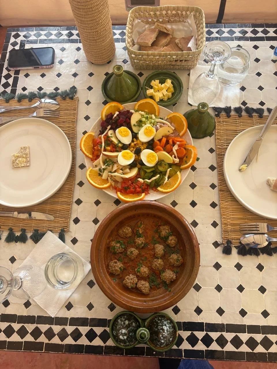A Moroccan-style meal setup on a table featuring a large bowl of salad with sliced oranges, boiled eggs, and vegetables, a terracotta dish with meatball stew, a basket of bread, small green bottles of oil, and two glasses of water. The table has wove
