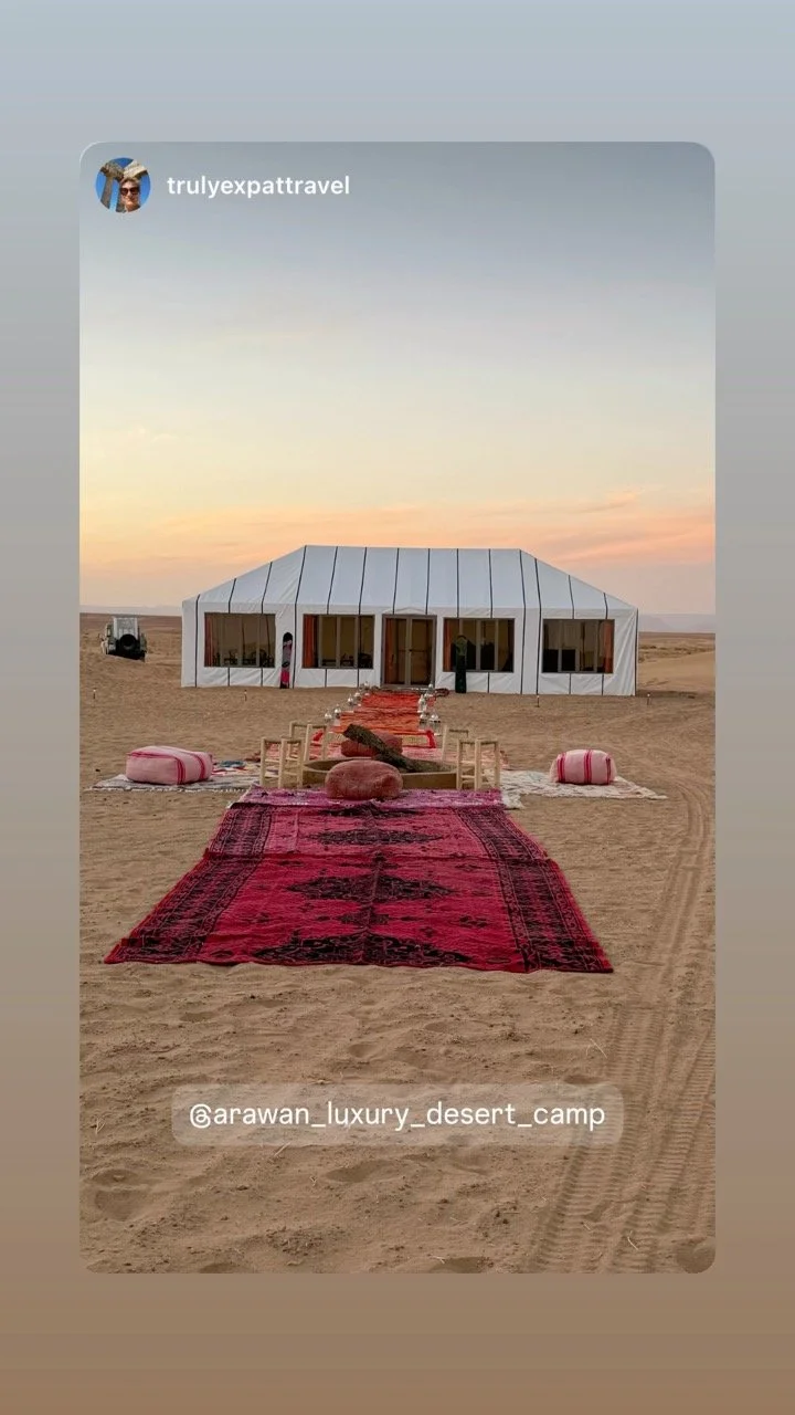Luxury desert camp with a large white tent, red carpets, and seating areas on sandy terrain at sunset.