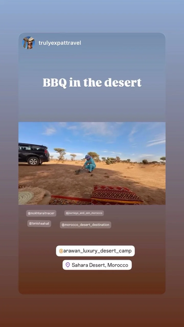 A person in blue clothing preparing a barbecue in the Sahara Desert, Morocco with a vehicle and desert trees in the background.