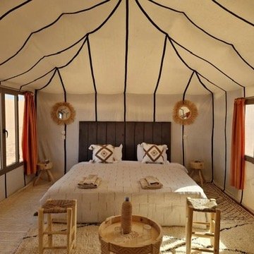 Interior of a bedroom in a canvas tent with a large bed, two pillows, and side tables, with orange curtains and decorative wreaths on the wall.