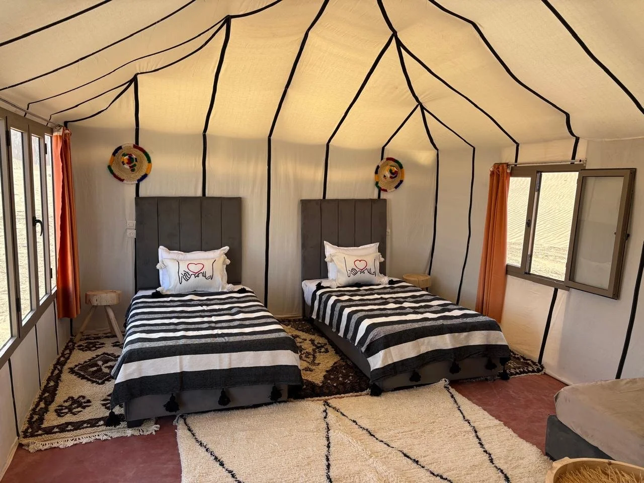 Two beds with black and white striped bedding and white pillows with heart designs are inside a tent with beige fabric walls and black lines creating a geometric pattern. Each bed has a headboard, and there are small side tables next to each bed. The