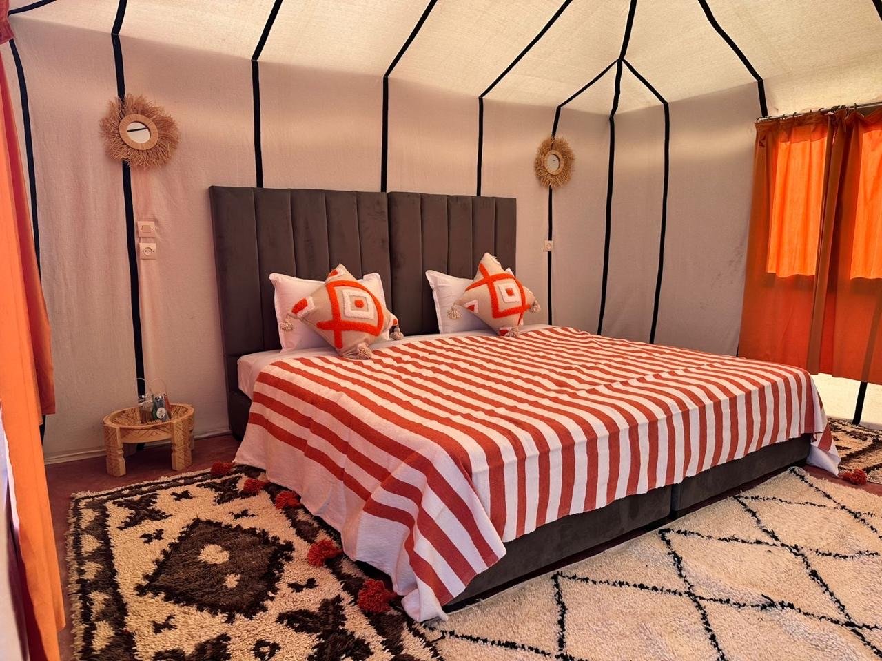 A bedroom with a large bed covered in a red and white striped bedspread, decorative pillows with a geometric pattern, a gray headboard, and a patterned rug. The walls are decorated with two round mirrors and orange curtains are hung on windows.