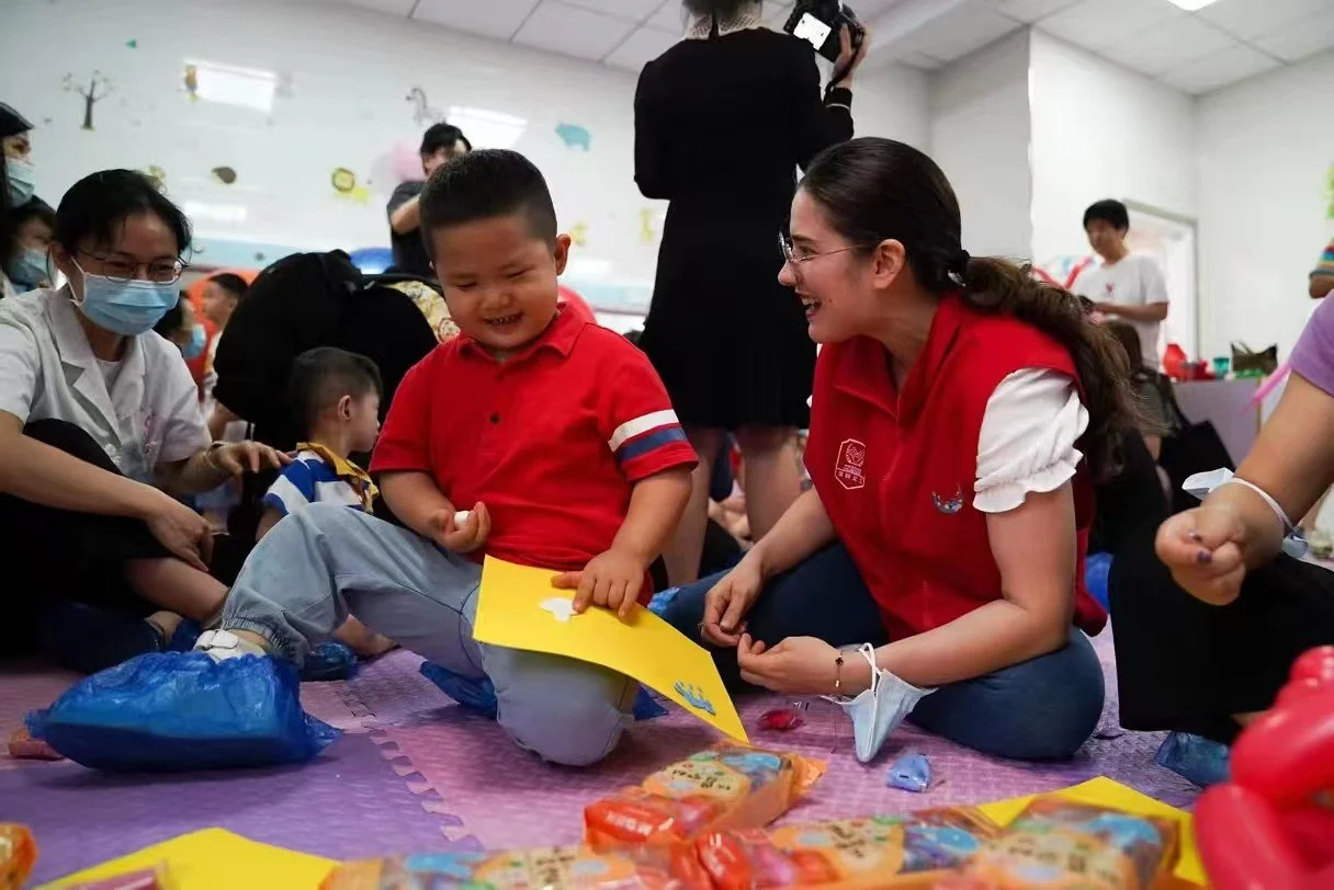 A group of children and adults sitting on a colorful mat indoors, engaging in a craft activity with paper and snacks. A woman in a red vest is smiling and talking with a young boy in a red shirt.