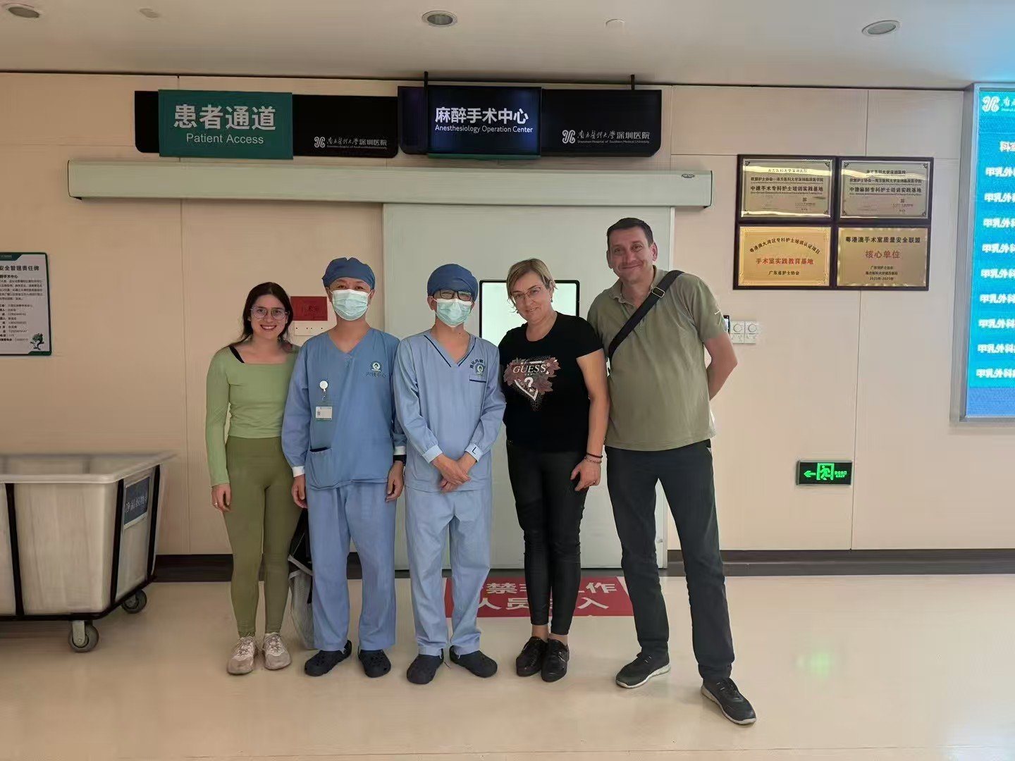 Group of five people standing in a hospital corridor, two women, two healthcare workers in scrubs and masks, and a man with a shoulder bag, behind signs indicating patient access and an anesthesia operation center.