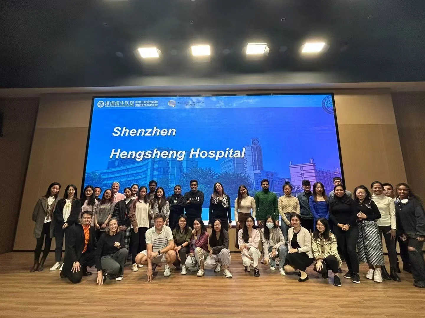 Group of people, including men and women of various ages and ethnicities, posing for a photo on a stage in front of a large digital screen displaying "Shenzhen Hengsheng Hospital." The stage has a wooden floor and is indoors.