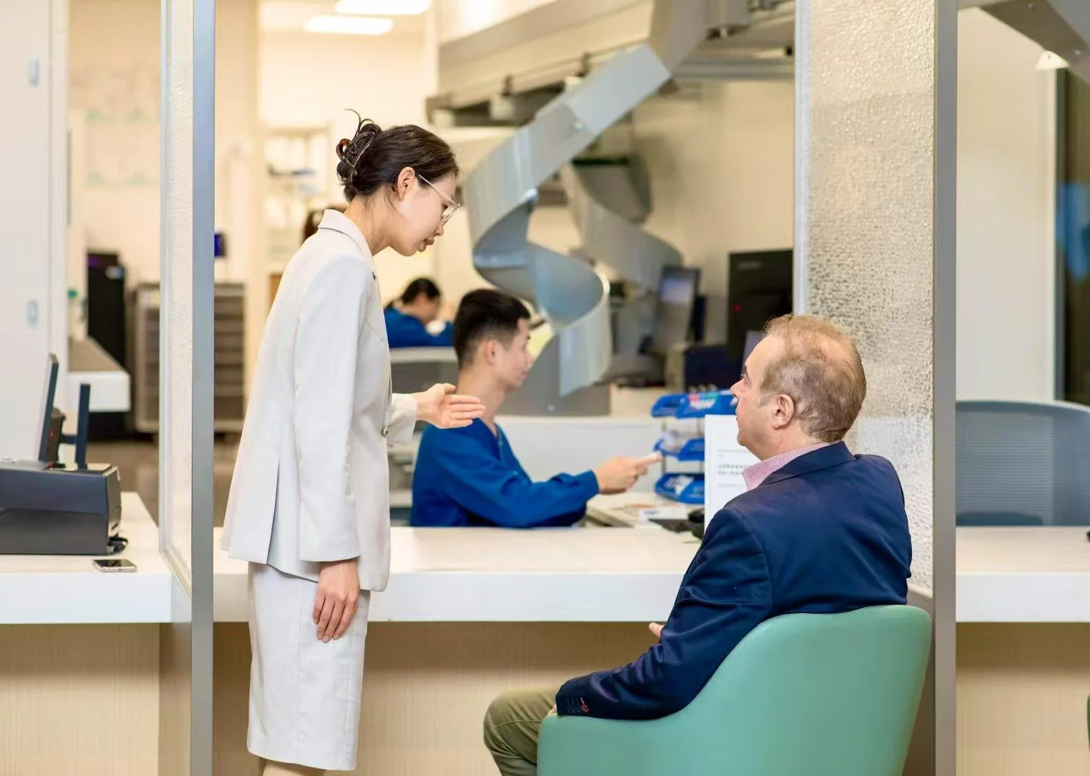 A woman in a beige suit talking to a man sitting in a lobby or reception area, with a healthcare or pharmacy setting behind her.