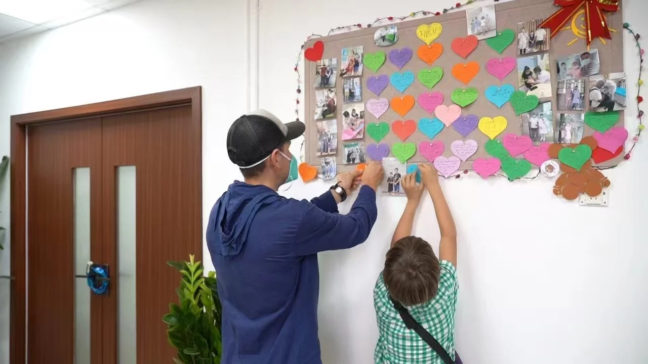 Adult and child placing colorful paper hearts on a wall display with photos and messages, decorated with string lights and a large bow, in a hallway.