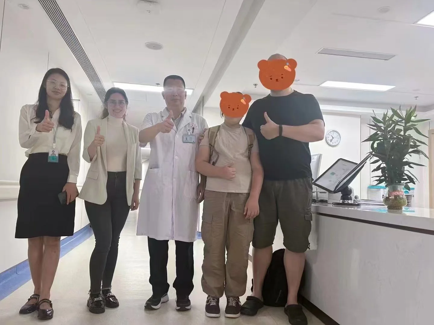 Group of five people standing in hospital corridor, three women, one man in a medical coat, and two persons with bear face stickers, some giving thumbs up.