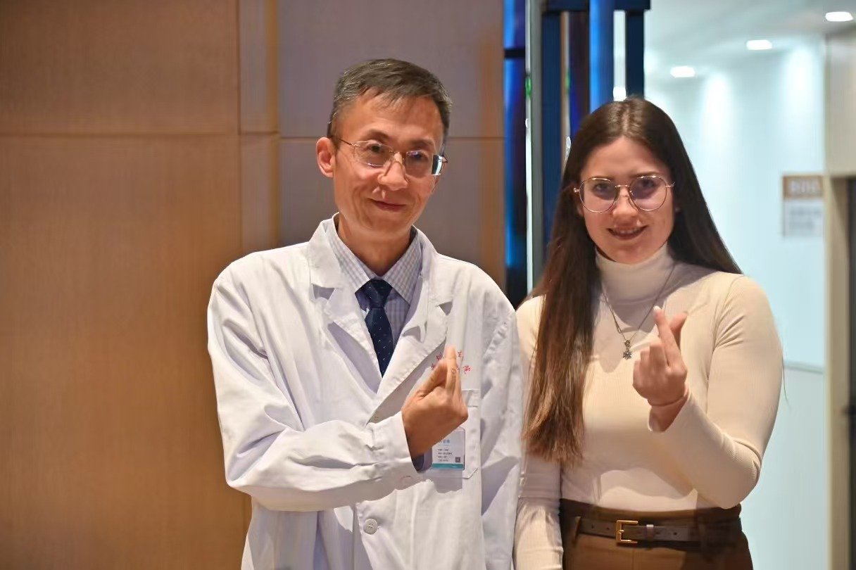 A man in a doctor's coat and a woman in glasses and a beige turtleneck making finger heart signs.