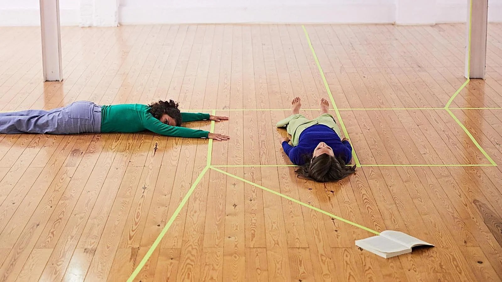 Two women lying on the wooden floor inside a room, separated by yellow tape, with an open book nearby.