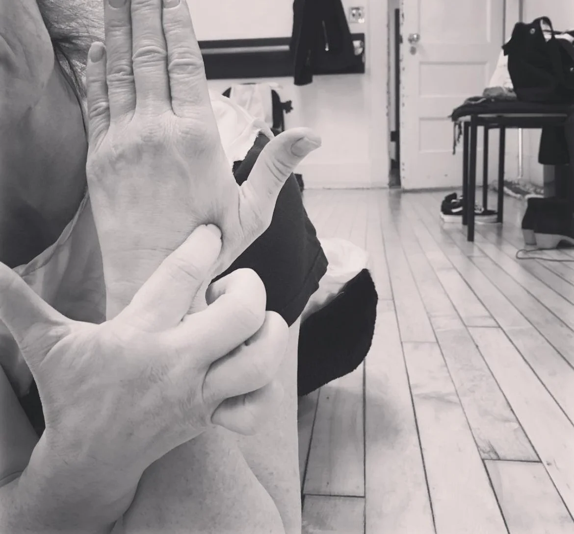 A person is seated indoors with their hand raised, showing the back of their hand and fingers. The photo is in black and white, with a room featuring a wooden floor, a door, a black chair, and some bags in the background.